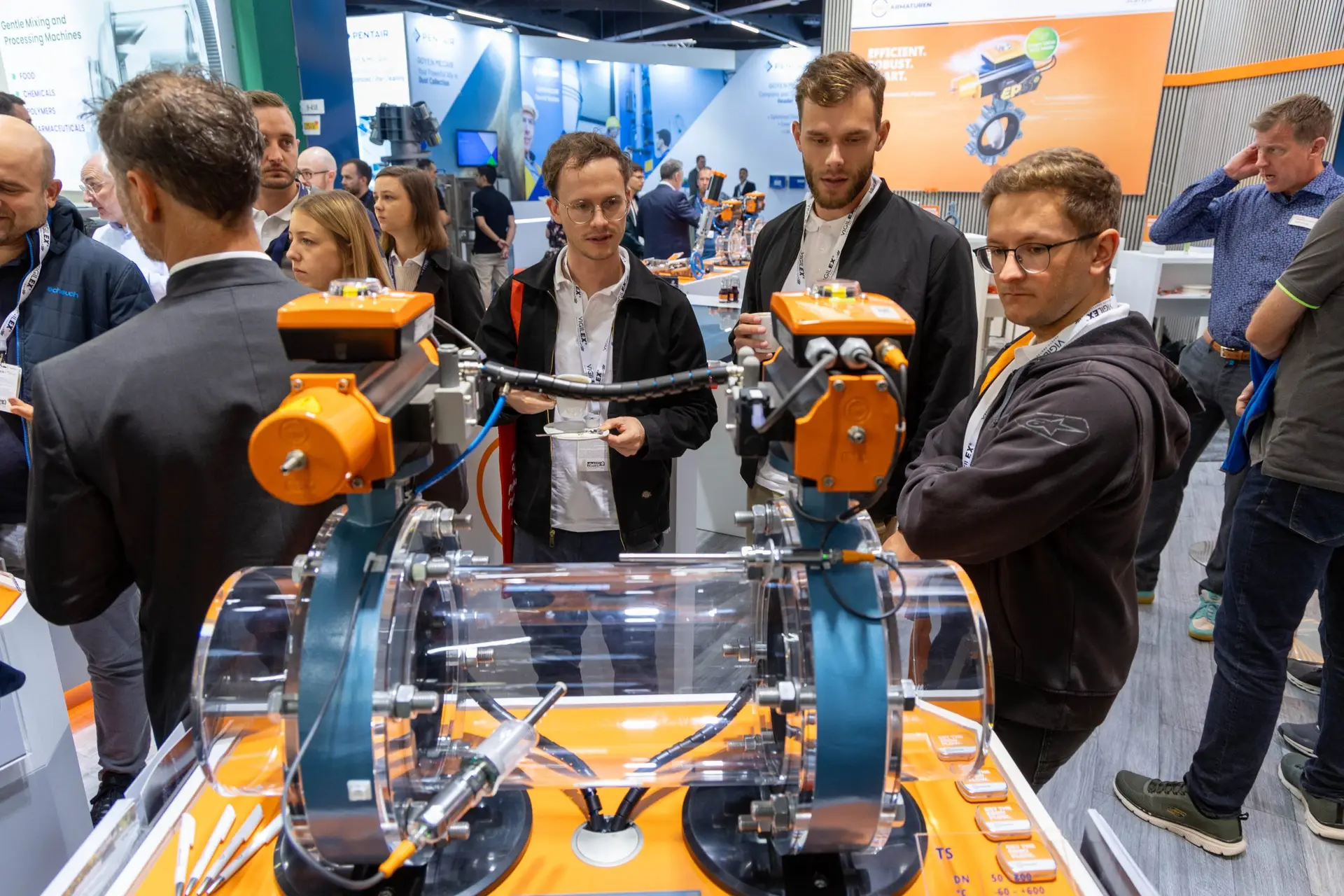 Fachbesucher betrachten auf der POWTECH TECHNOPHARM Industriearmaturen mit pneumatischen Antrieben an einem transparenten Rohrleitungsmodell