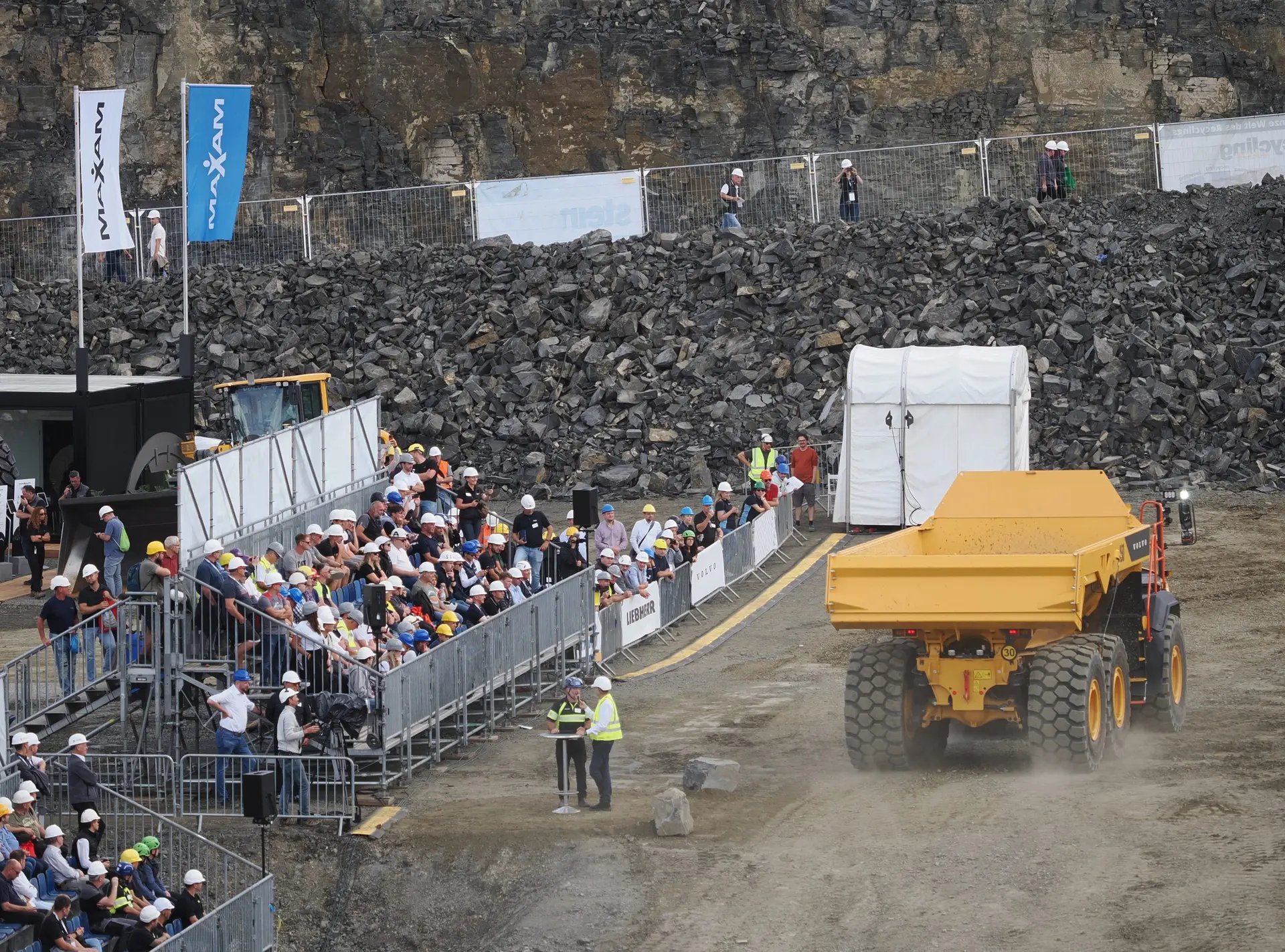 Zuschauer auf einer Tribüne verfolgen die Live-Demonstration eines gelben knickgelenkten Muldenkippers im Steinbruch der steinexpo