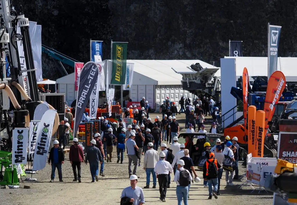 Besucher mit Schutzhelmen zwischen Ausstellerständen und Beachflags auf dem steinexpo-Gelände, im Hintergrund die Steinbruchwand