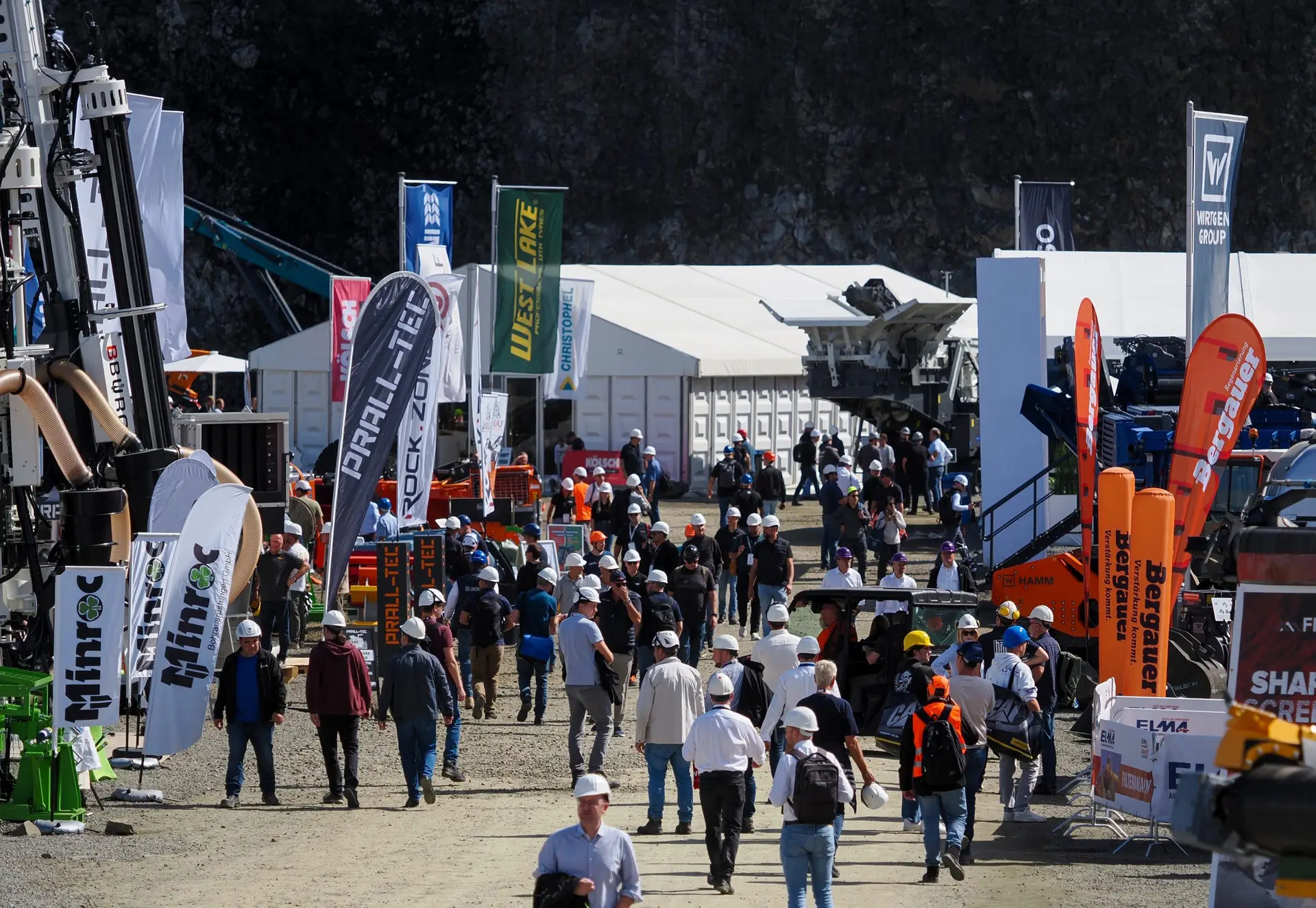 Besucher mit Schutzhelmen zwischen Ausstellerständen und Beachflags auf dem steinexpo-Gelände, im Hintergrund die Steinbruchwand