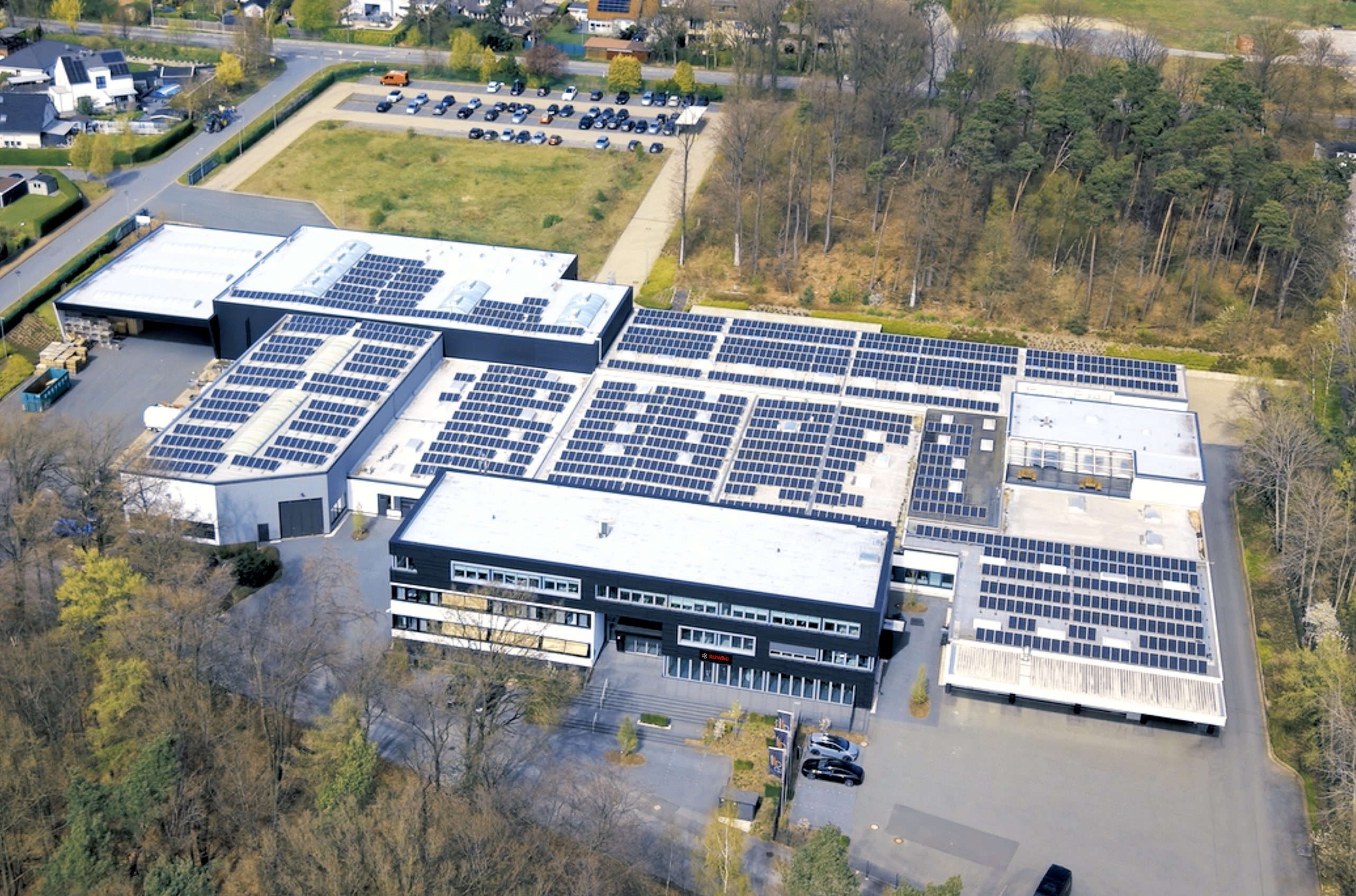 Luftaufnahme eines Industrie- und Verwaltungsstandorts mit Produktionsgebäuden, Parkflächen und Photovoltaikanlagen auf den Dachflächen.