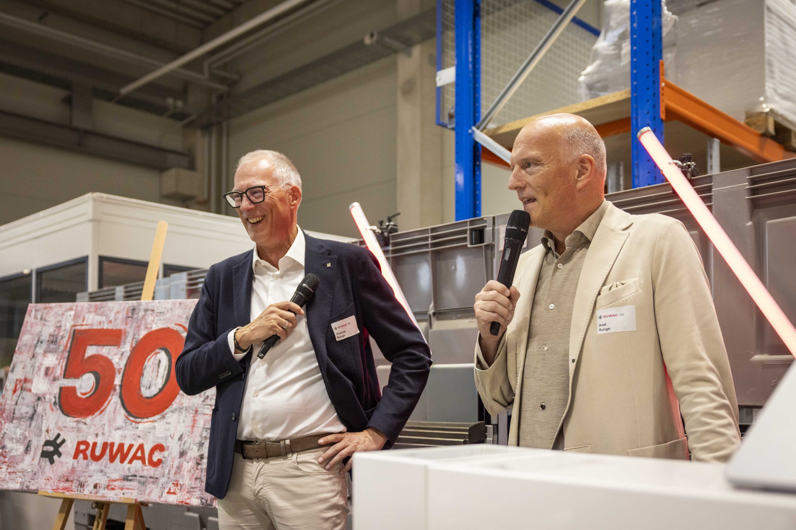 Zwei Unternehmensleiter sprechen während einer Jubiläumsveranstaltung vor Mitarbeitenden in einer Produktionshalle.