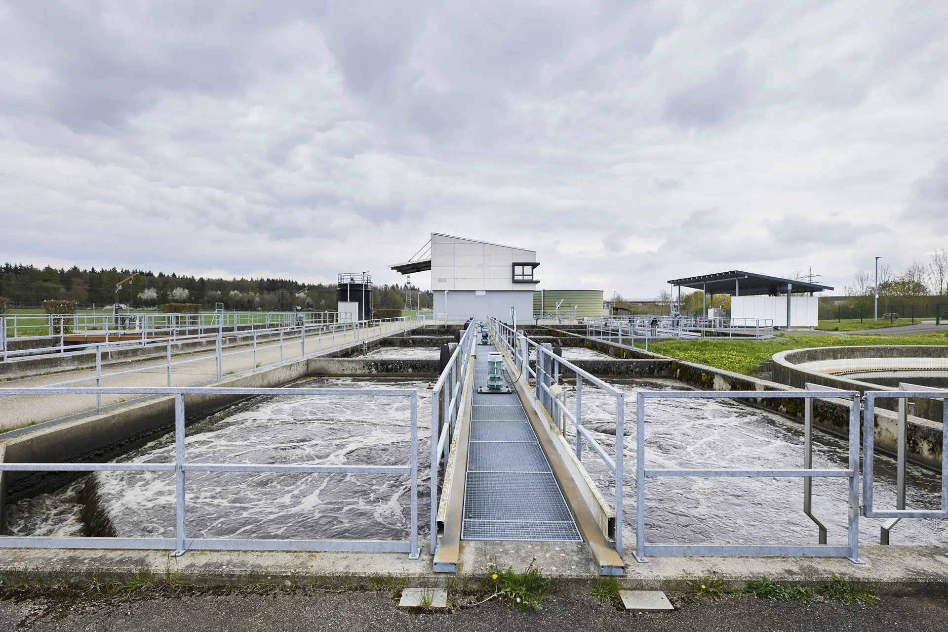 Belüftete Becken mit aufgewühlter Wasseroberfläche, getrennt durch einen Steg mit Geländer, im Hintergrund ein Betriebsgebäude und ein rundes Klärbecken