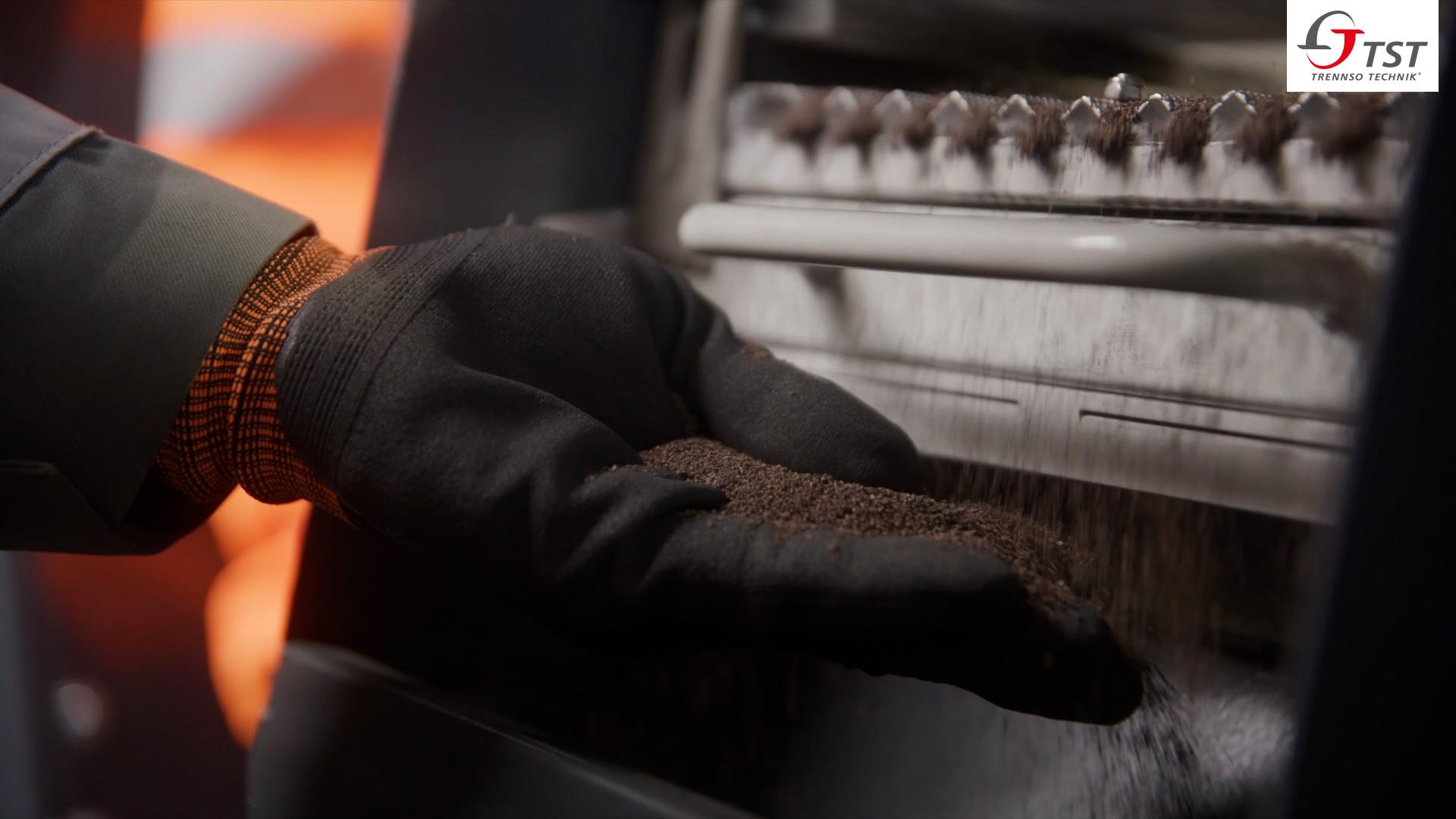 Behandschuhte Hand prüft feinkörniges dunkles Recyclingmaterial, das aus einer Maschine herabrieselt