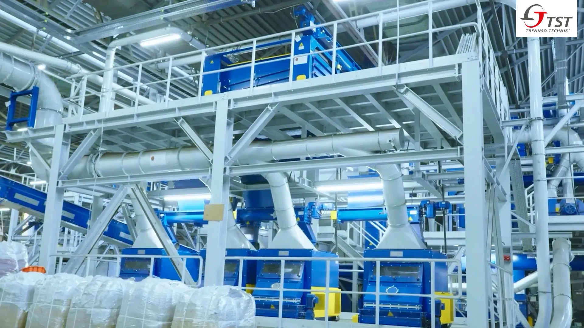 Mehrstöckige Aufbereitungsanlage in einer Halle mit blauen Maschinen auf Stahlbühnen, Absaugleitungen und Förderbändern, davor Big Bags